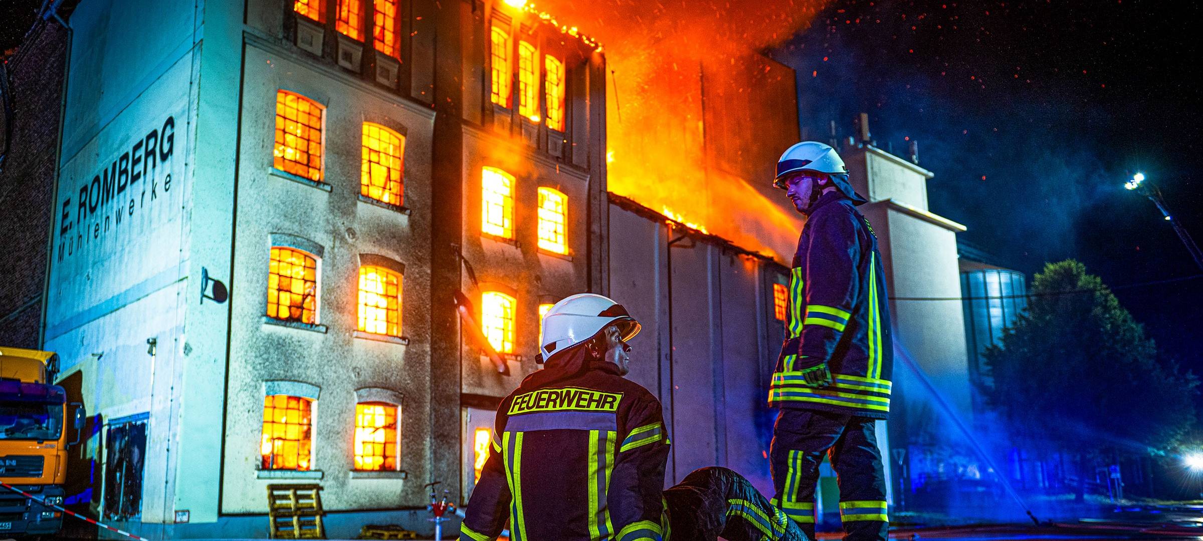 Zwei Feuerwehrleute vor einem brennenden Gebäude. Der eine sitzt erschöpft der andere steht.