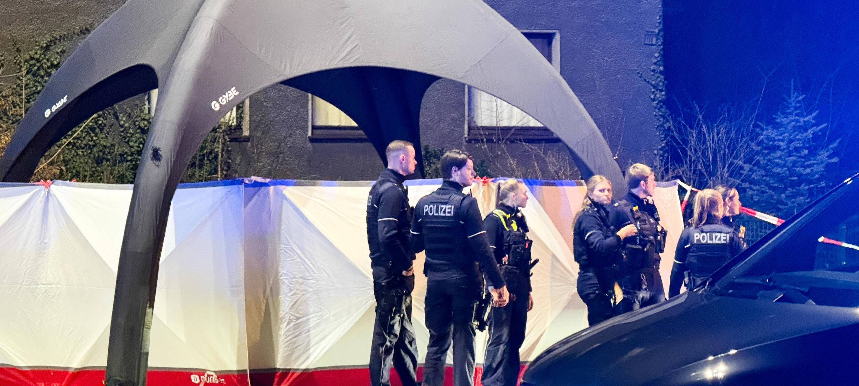 Polizisten stehen vor dem Tatort, der durch einen weißen Sichtschutz und einem grauem Zelt abgedeckt ist.
