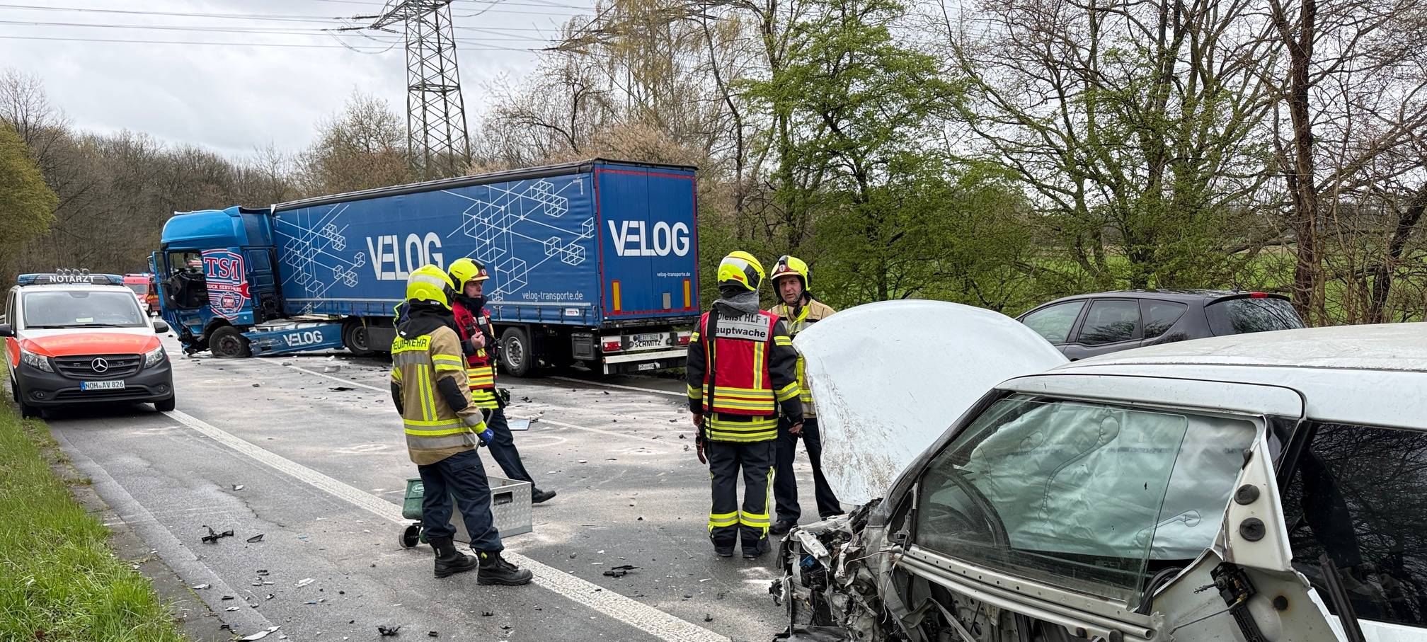 Das stark beschädigte Auto ist im Vordergrund zu sehen. Rettungskräfte stehen davor. Im Hintergrund ist der beschädigte LKW zu sehen.