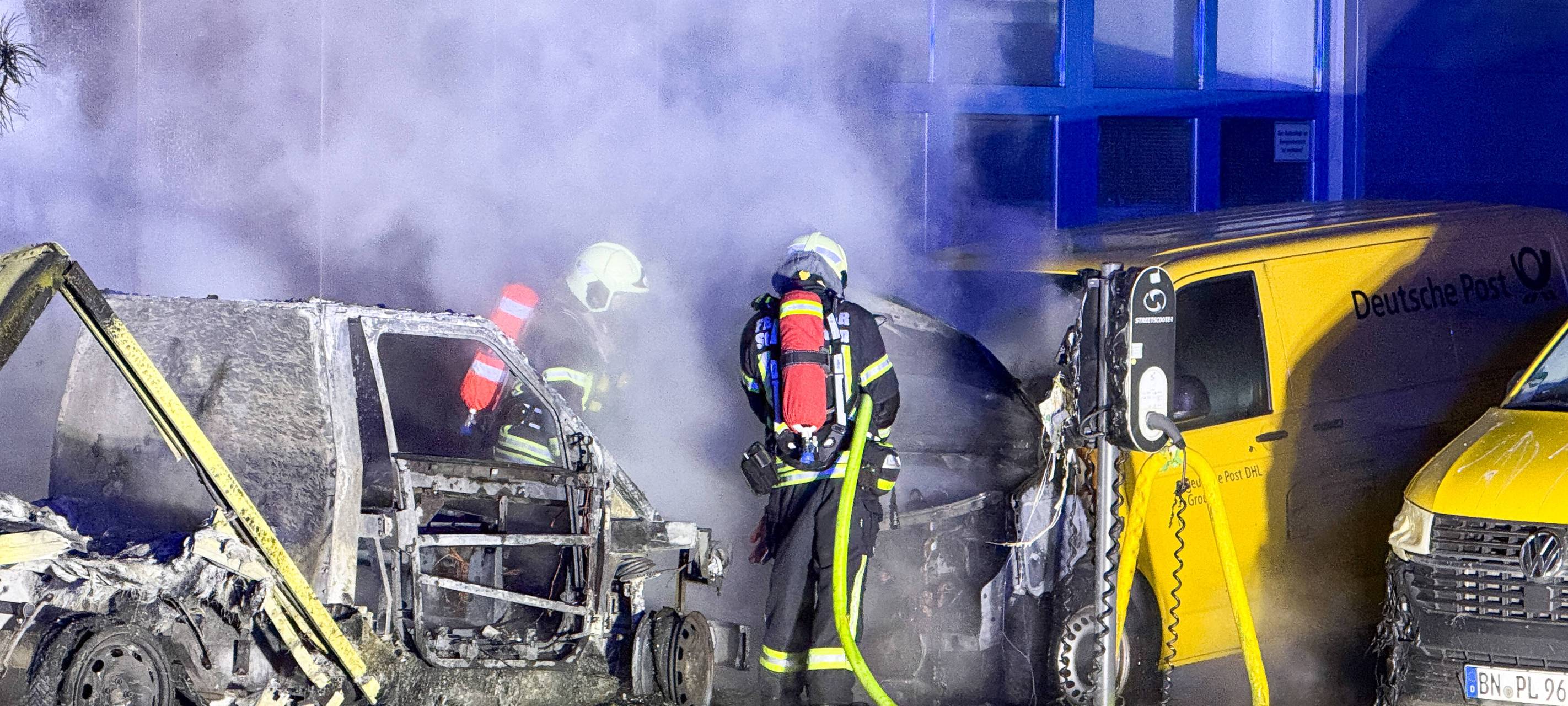 Postfahrzeuge sind ausgebrannt. Einsatzkräfte der Feuerwehr stehen für Nachlöscharbeiten an den qualmenden Autos.